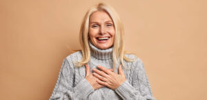Cheerful good looking middle aged woman keeps hands pressed on chest smiles broadly and expresses positive emotions dressed in winter jumper happy to hear compliment isolated over beige background